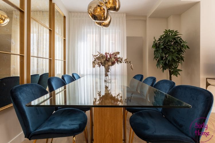 Comedor reformado con mesa de cristal sobre pie de madera, sillas de terciopelo azul y pared de espejos con perfil dorado