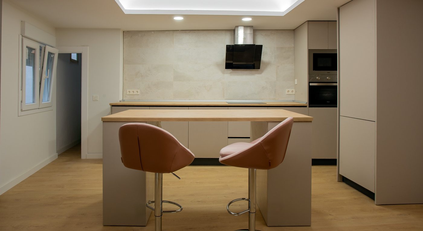 Cocina con isla central de sobre de madera, dos taburetes y techo con luz indirecta
