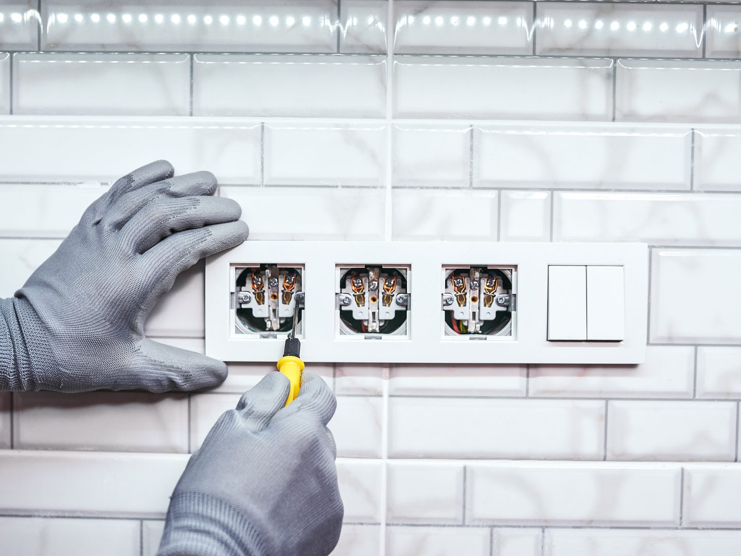 Electricista con guantes conectando tres enchufes y un interruptor en una pared alicatada