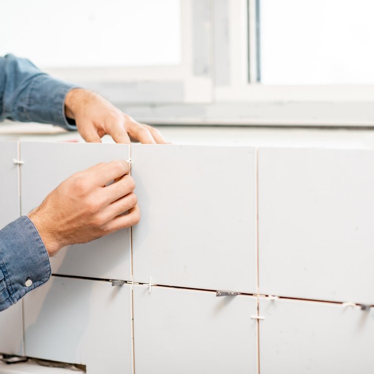 Manos colocando azulejo blanco con crucetas en la pared del baño