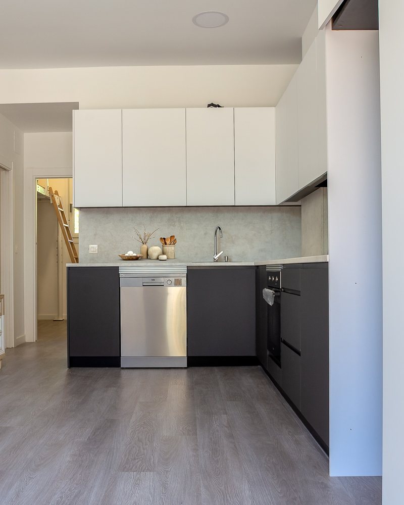 Cocina abierta tras la reforma, con muebles altos blancos, bajos grafito y suelo de lamas
