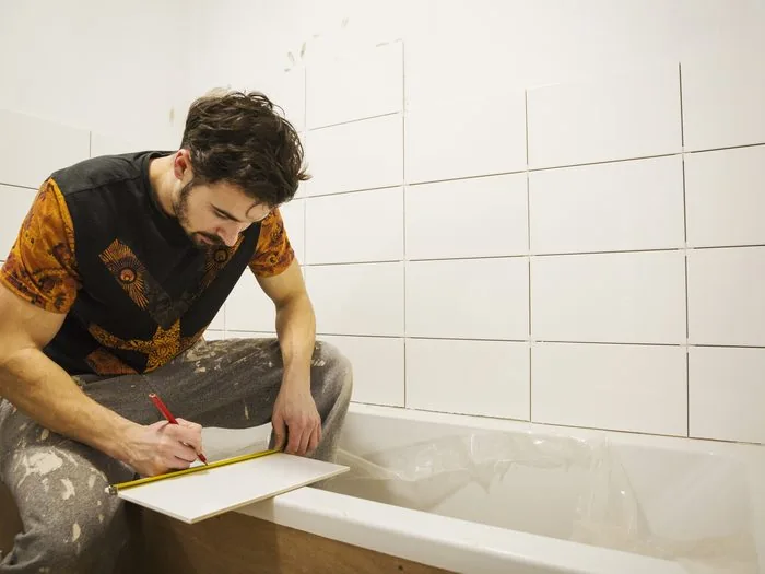 Albañil sentado en el borde de la bañera marcando con lápiz un azulejo blanco para cortarlo, cuánto cobra un albañil Zaragoza