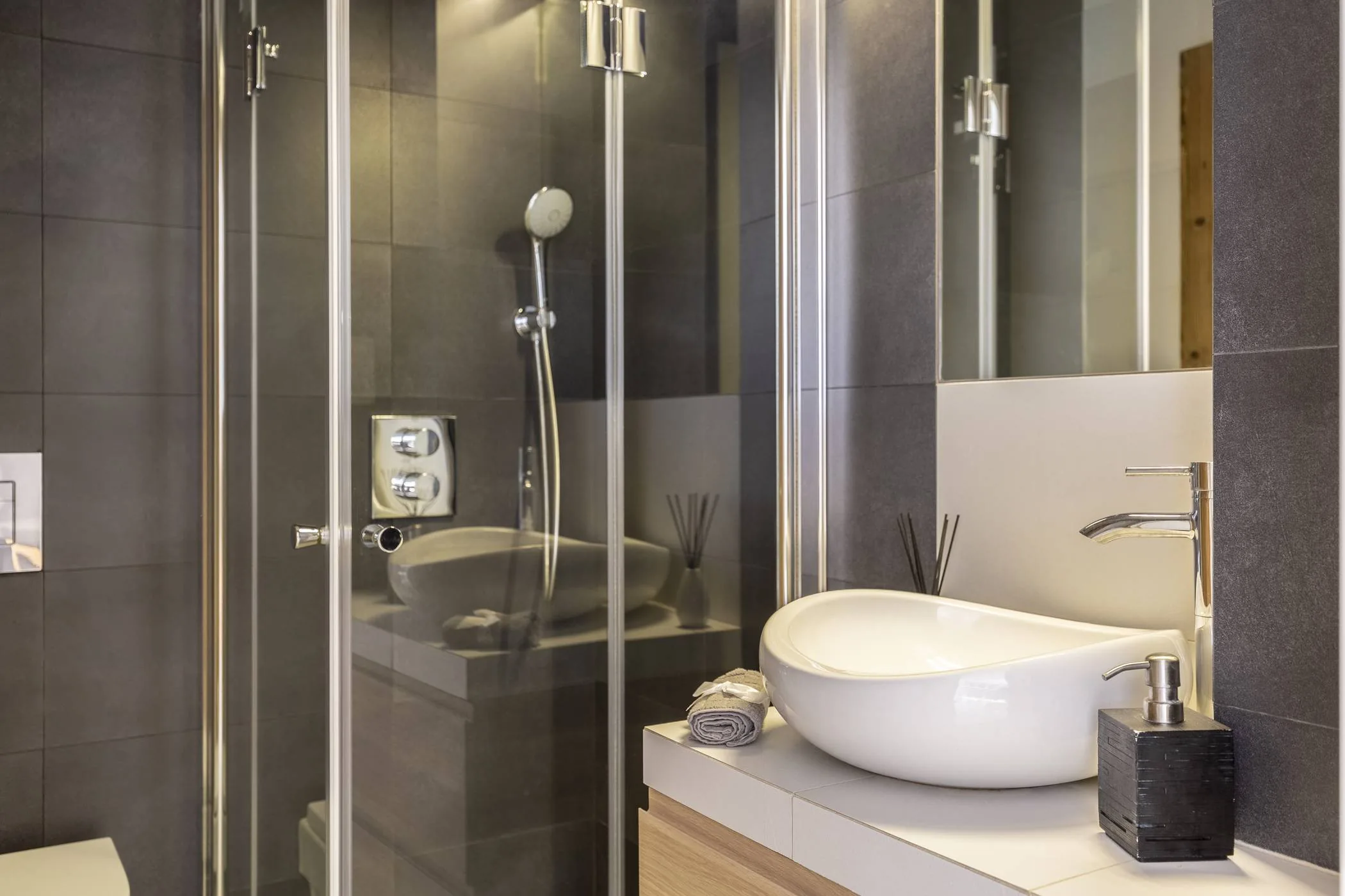 Baño pequeño con ducha de mampara de vidrio, lavabo sobre encimera y azulejo gris oscuro, precio reforma de baño Zaragoza