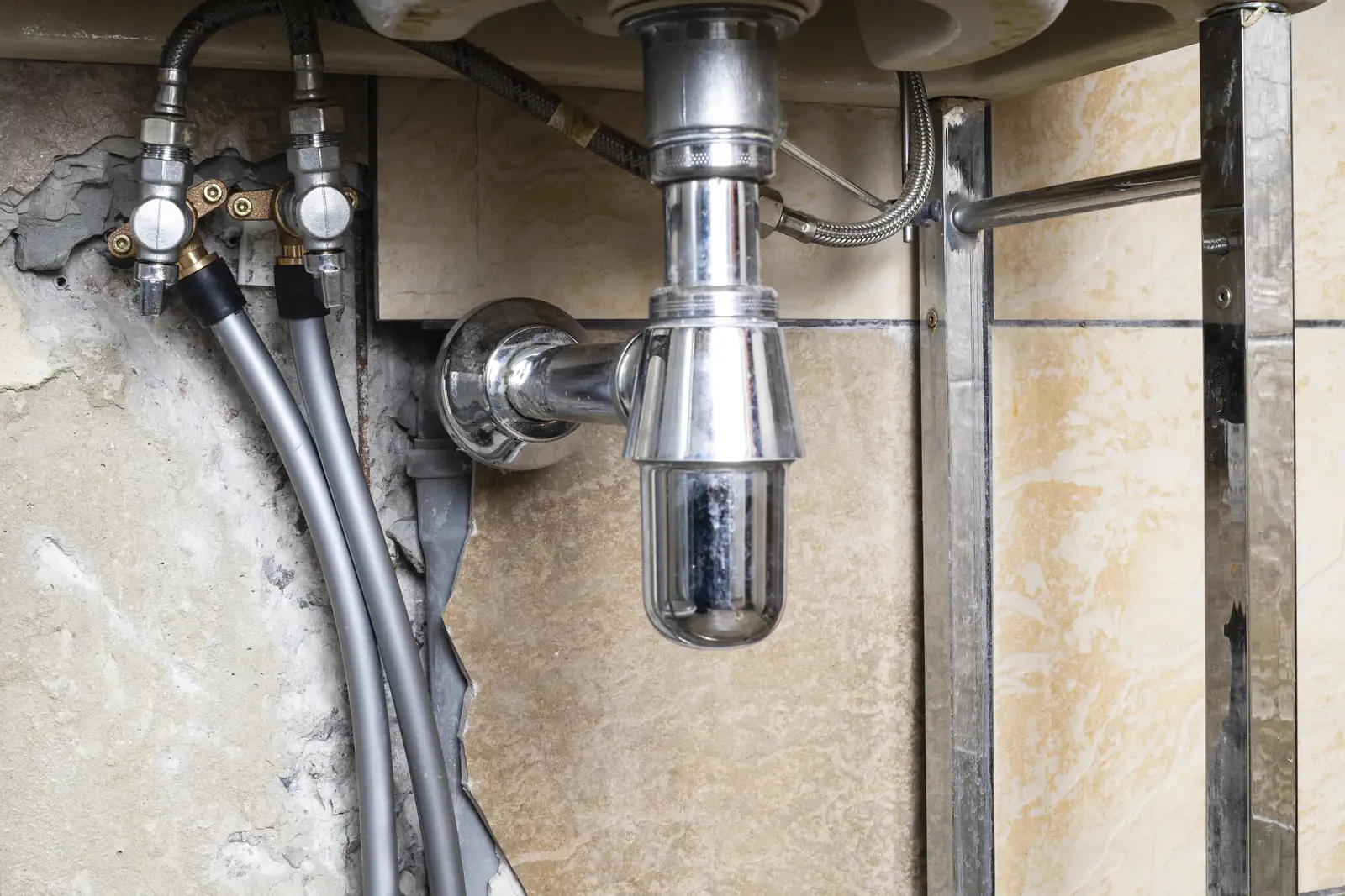 Sifón cromado y latiguillos bajo un lavabo, con las llaves de paso y los tubos saliendo de la pared abierta, precio reparación fuga de agua Zaragoza